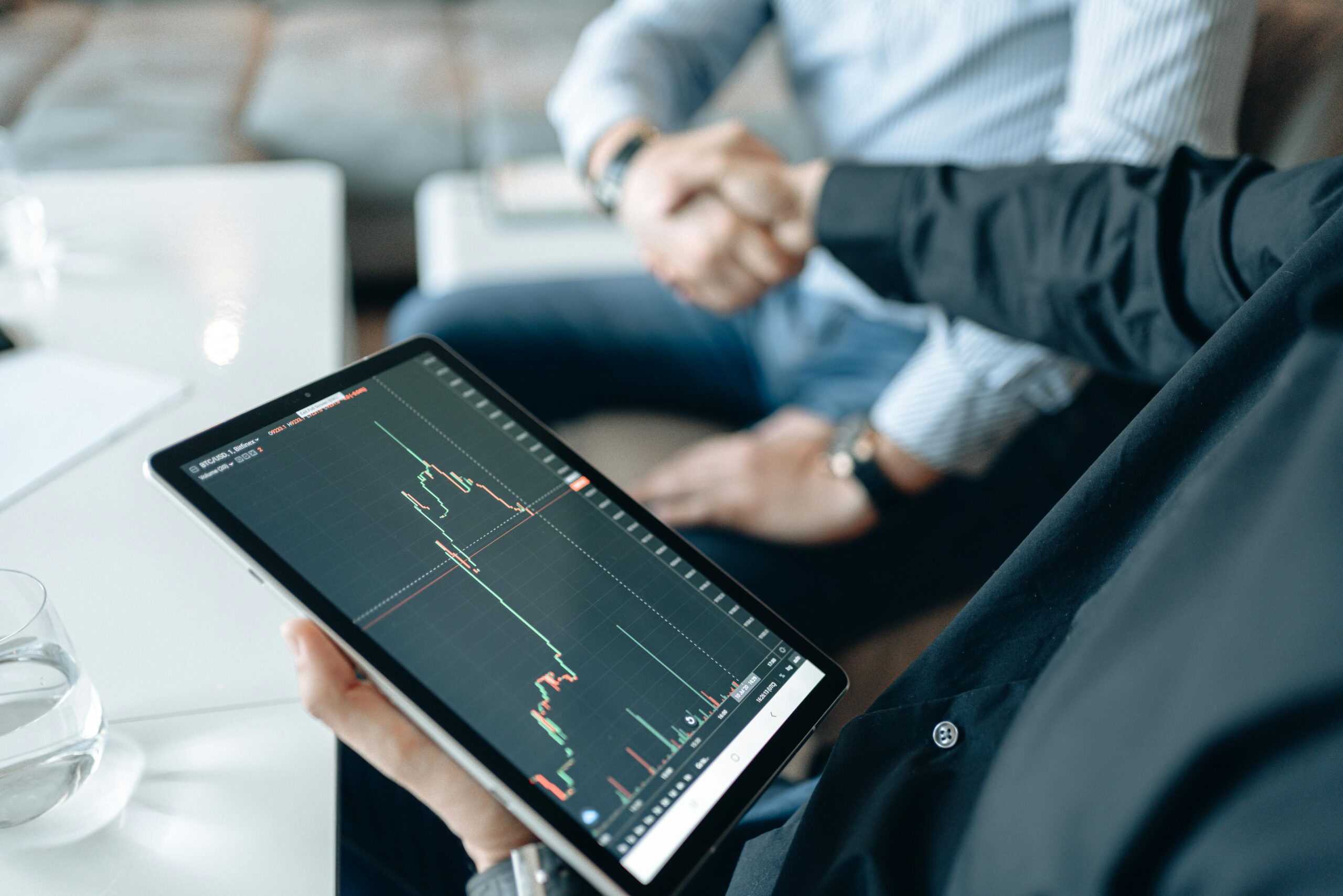 Two adults shaking hands with a stock market chart on a tablet, symbolizing a business deal.