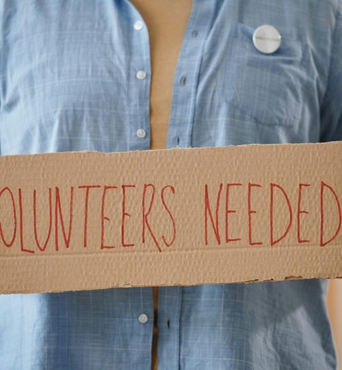 Person holding a sign reading 'Volunteers Needed' to encourage community support.