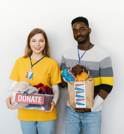 Two diverse volunteers smiling while holding donation boxes indoors with a white background.